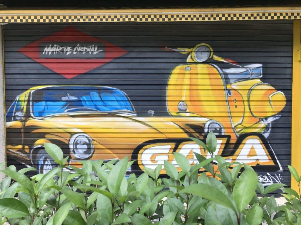 Graffiti en cierre metálico de GALA Autoescuela con coche clásico amarillo y moto Vespa