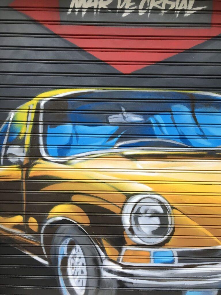 Detalle lateral del coche clásico amarillo pintado sobre cierre metálico de GALA Autoescuela