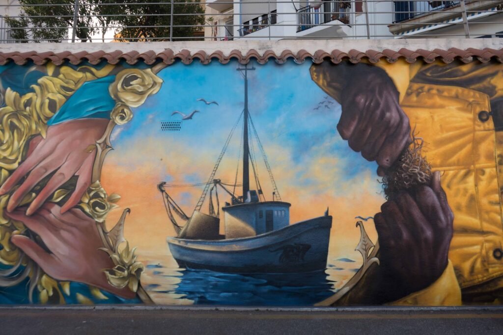 Barca de pesca al amanecer enmarcada por las manos de las dos figuras del mural de l'Ametlla de Mar