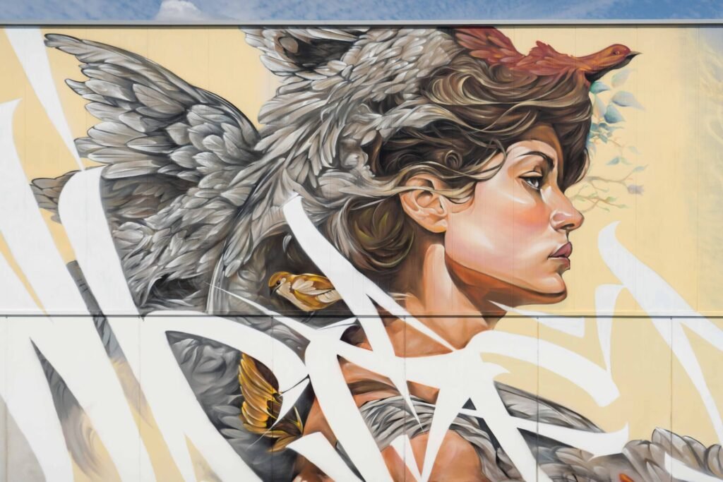 Mural Lliure inspirado en Niké: rostro de mujer entre alas grises y pájaros sobre fondo amarillo crema