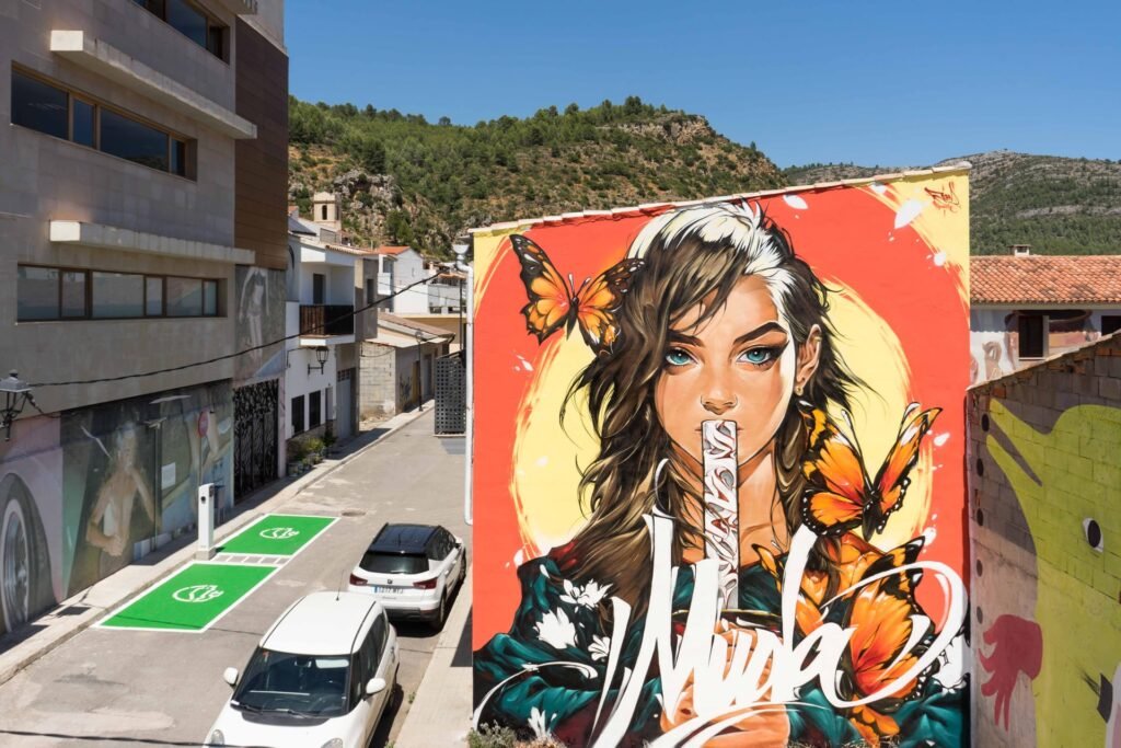 Mural Muda en Fanzara: mujer de ojos turquesa con mariposas y lettering blanco en una medianera del MIAU 2026