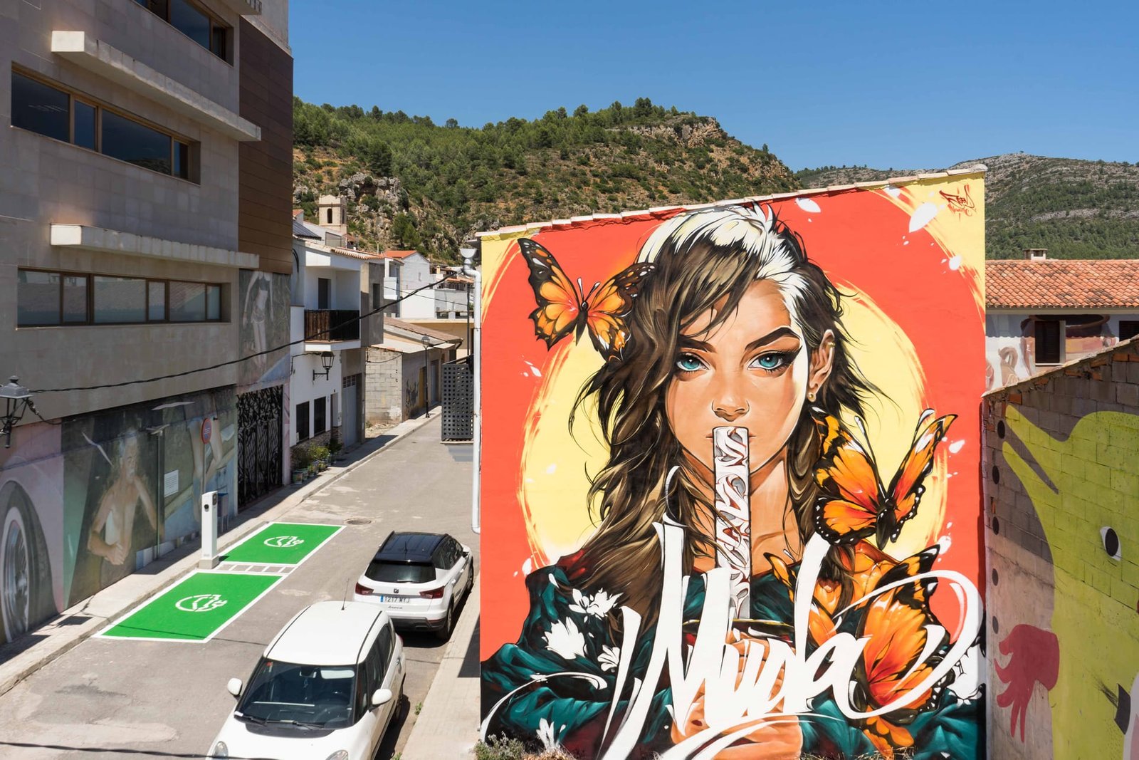 Mural Muda en Fanzara: mujer de ojos turquesa con mariposas y lettering blanco en una medianera del MIAU 2026